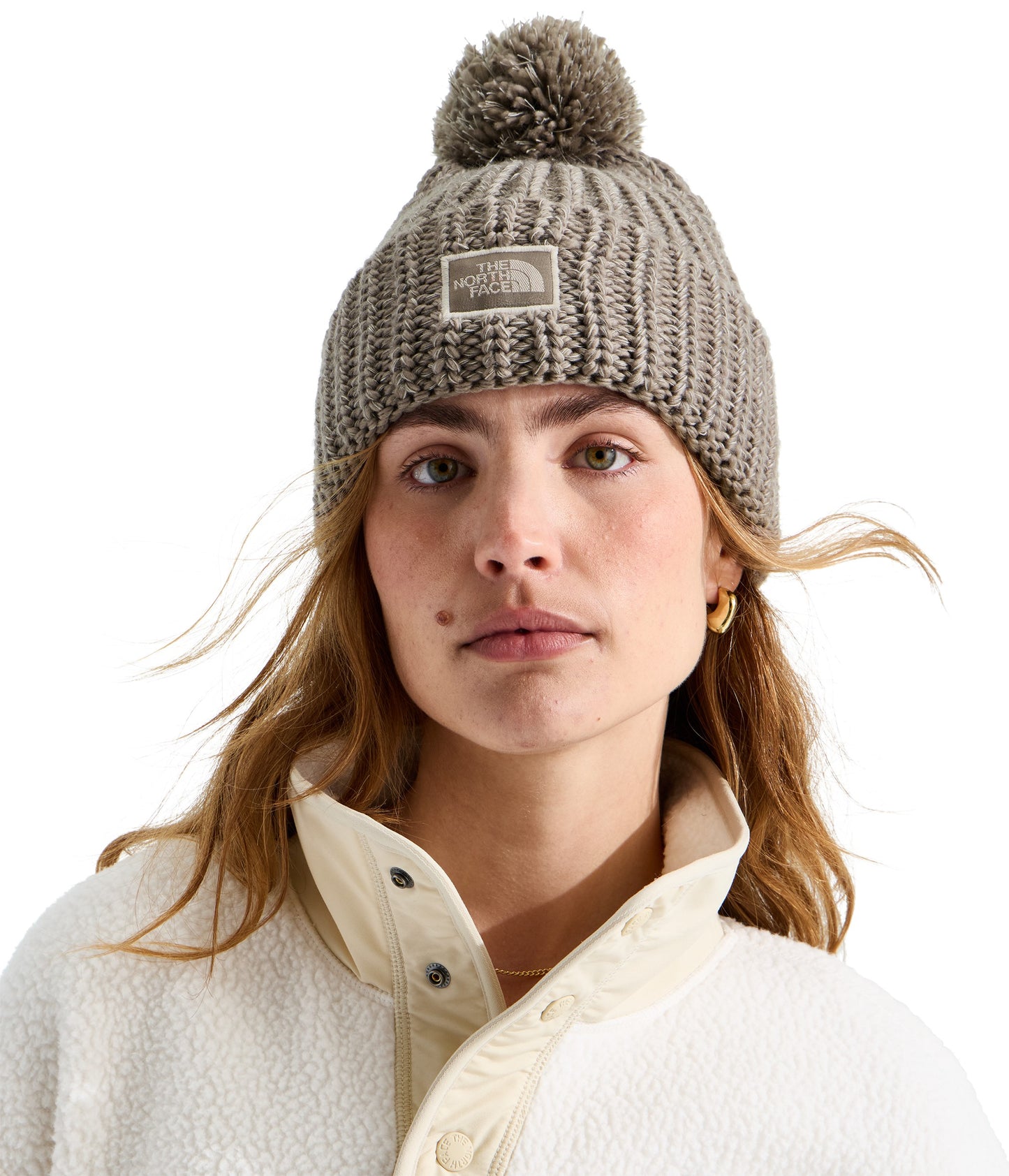 A woman wearing a taupe-colored, ribbed-knit beanie with a matching pom-pom on top and a rectangular embroidered logo patch on the front, paired with a white, button-up sherpa fleece jacket.