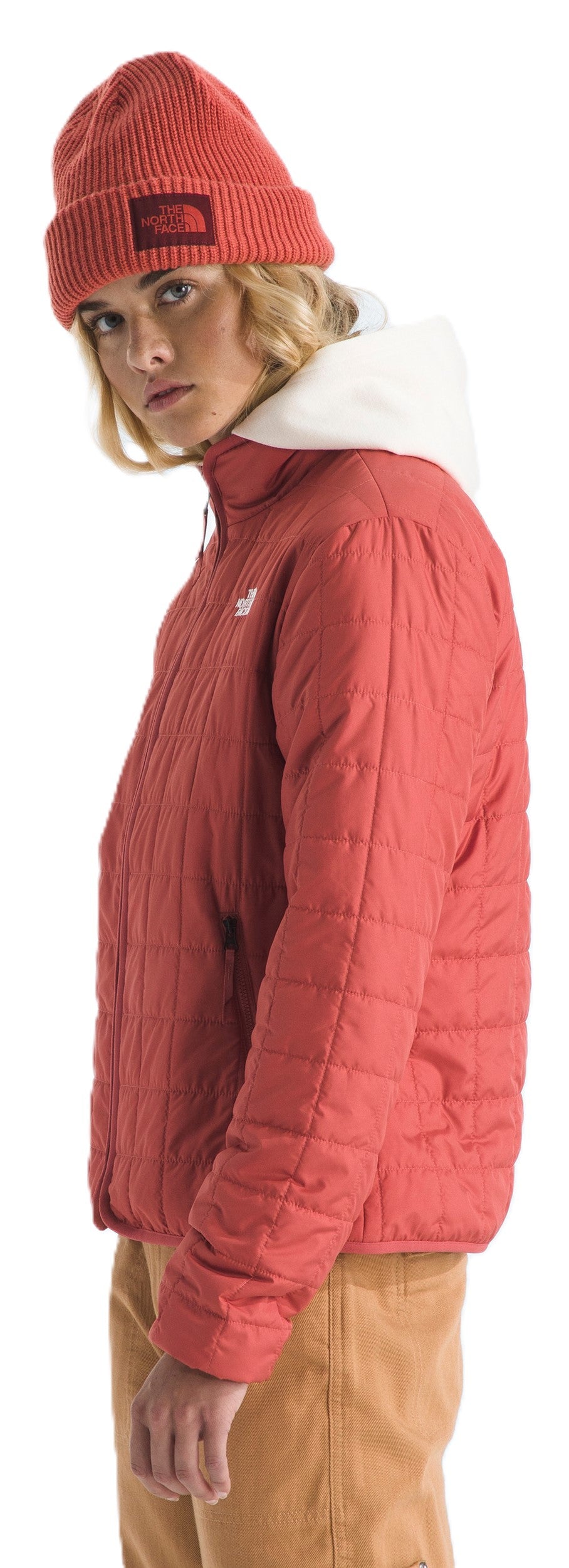 A person wearing a red knit hat and quilted jacket, along with tan pants, in a studio shot.