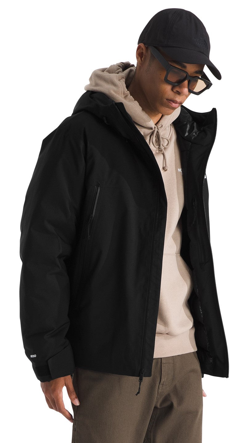 A man wearing a black hooded jacket with velcro cuffs over a beige hoodie, brown pants, black-framed glasses, and a black baseball cap.