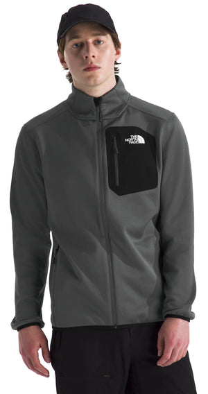 A man wearing a dark grey, zip-up fleece jacket with a black chest pocket and a white logo on the chest pocket, paired with a black baseball cap and black pants.