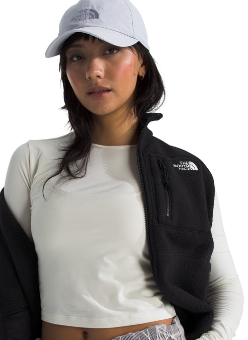 A woman wearing a light-colored baseball cap, a white long-sleeved shirt, and a black fleece vest with a small zippered chest pocket featuring a logo.