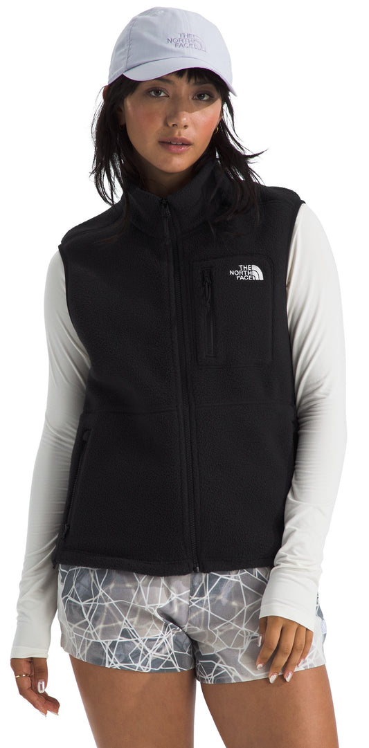 A person wears a light grey baseball cap, a black zippered fleece vest with a small white logo on the chest, a long-sleeved light-colored athletic shirt, and patterned grey and white running shorts.