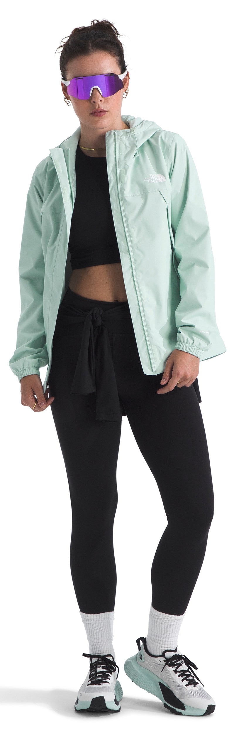 A woman wearing a light green jacket, black sports bra, black shorts, black leggings, white socks and sneakers, topped off with futuristic purple sunglasses.