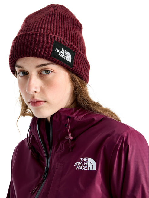 A young woman wearing a ribbed, burgundy beanie with a rectangular black logo patch on the front, paired with a matching burgundy windbreaker jacket that has a white logo printed on the chest.