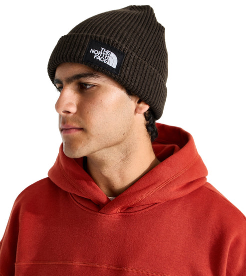 A man wearing a dark olive ribbed knit beanie with a square black and white label featuring the text THE NORTH FACE, worn over a plain red hooded sweatshirt.