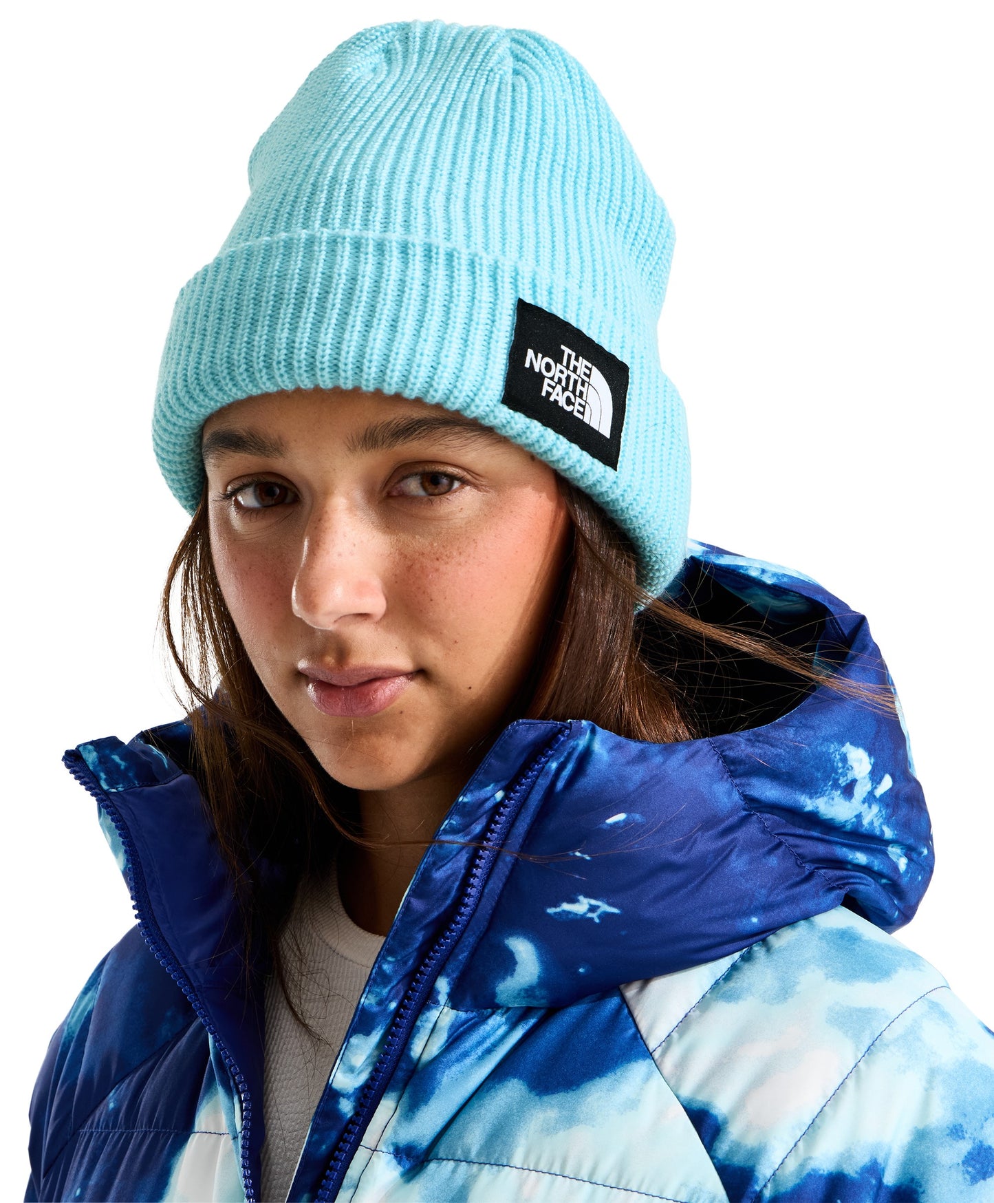 A young woman wearing a light blue ribbed knit beanie with a rectangular black label featuring white text, paired with a blue and white tie-dye hooded jacket.