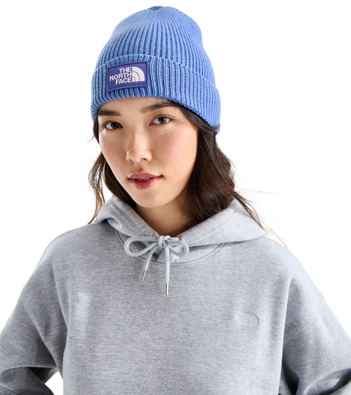 A woman wearing a light blue ribbed-knit beanie with a rectangular purple patch on the front displaying white text that reads THE NORTH FACE, over a light grey hooded sweatshirt.
