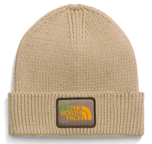 A tan knit beanie with a folded cuff featuring a brown rectangular patch with yellow text reading THE NORTH FACE and a small green graphic icon.