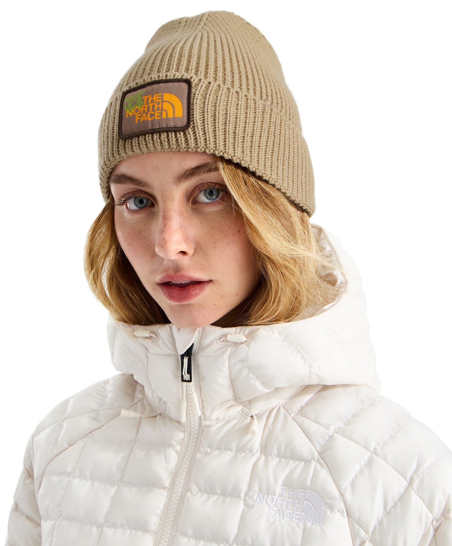 A fair-skinned woman with blonde hair wears a tan-colored ribbed beanie with a square brown and yellow patch that says The North Face on the front, paired with a white, quilted, hooded jacket.