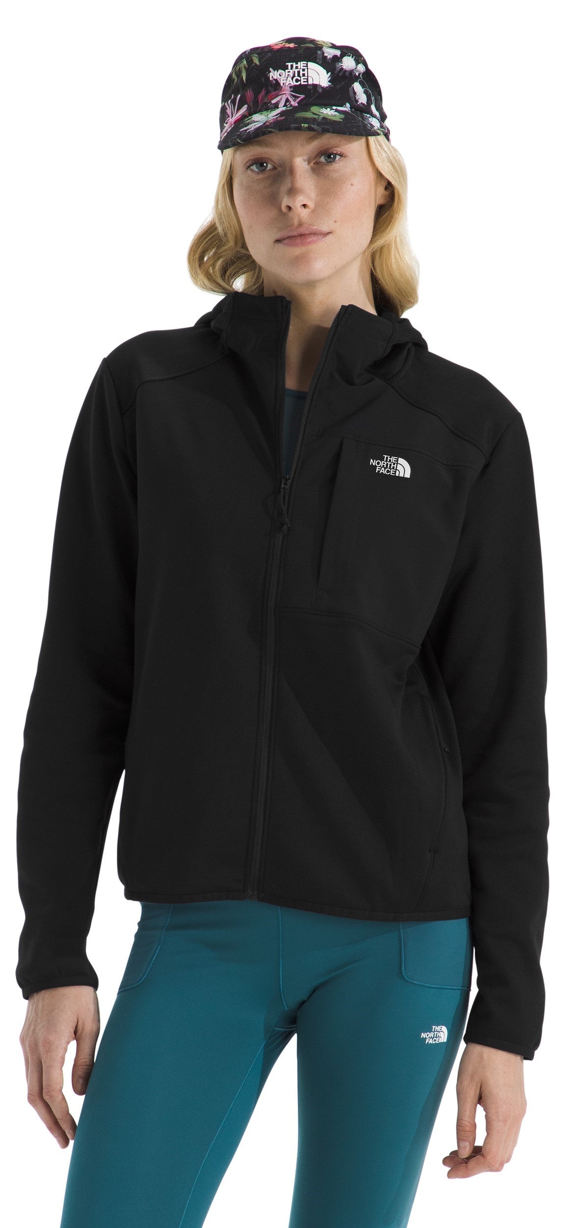 A woman wearing a black zip-up hoodie with a small logo on the left chest, a floral-patterned baseball cap, and teal leggings.