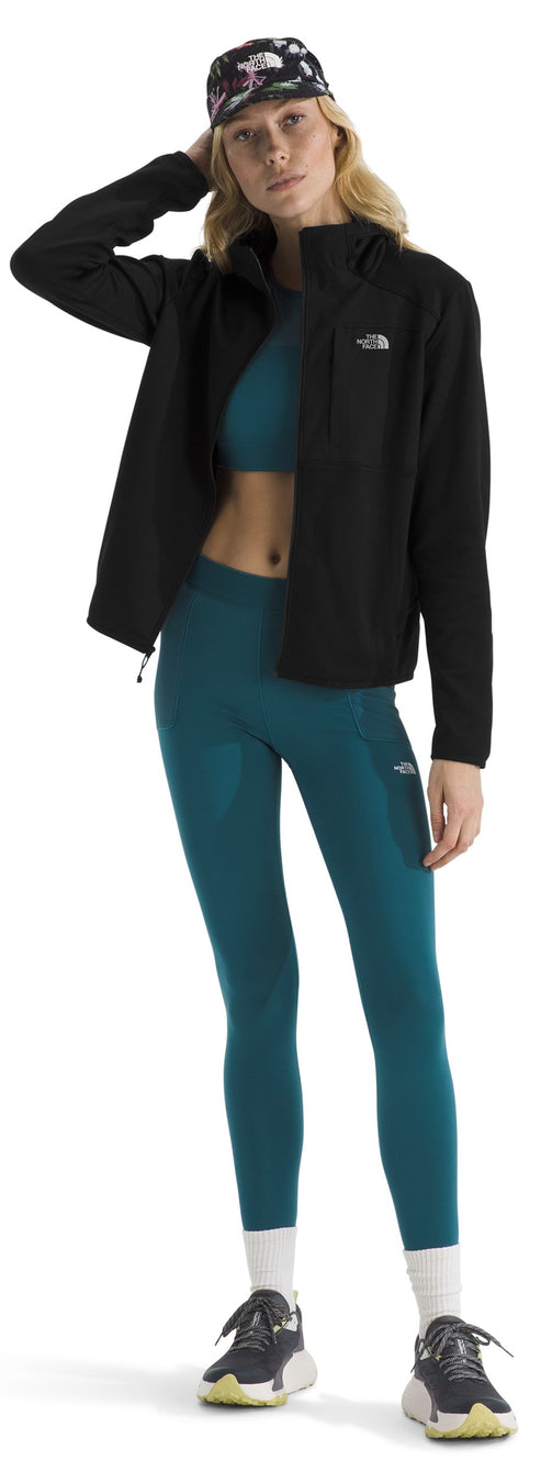 A woman stands posing in a black zip-up jacket, dark teal sports bra, and matching teal high-waisted leggings, paired with white crew socks and grey and yellow sneakers, while wearing a dark floral-patterned baseball cap.