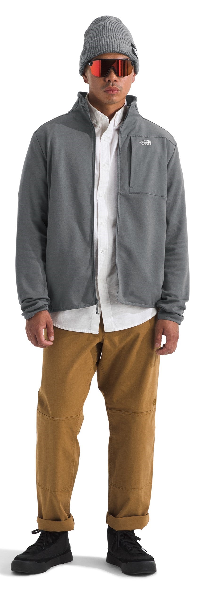 A person in a grey jacket, white shirt, and brown pants wearing a grey beanie and orange goggles.
