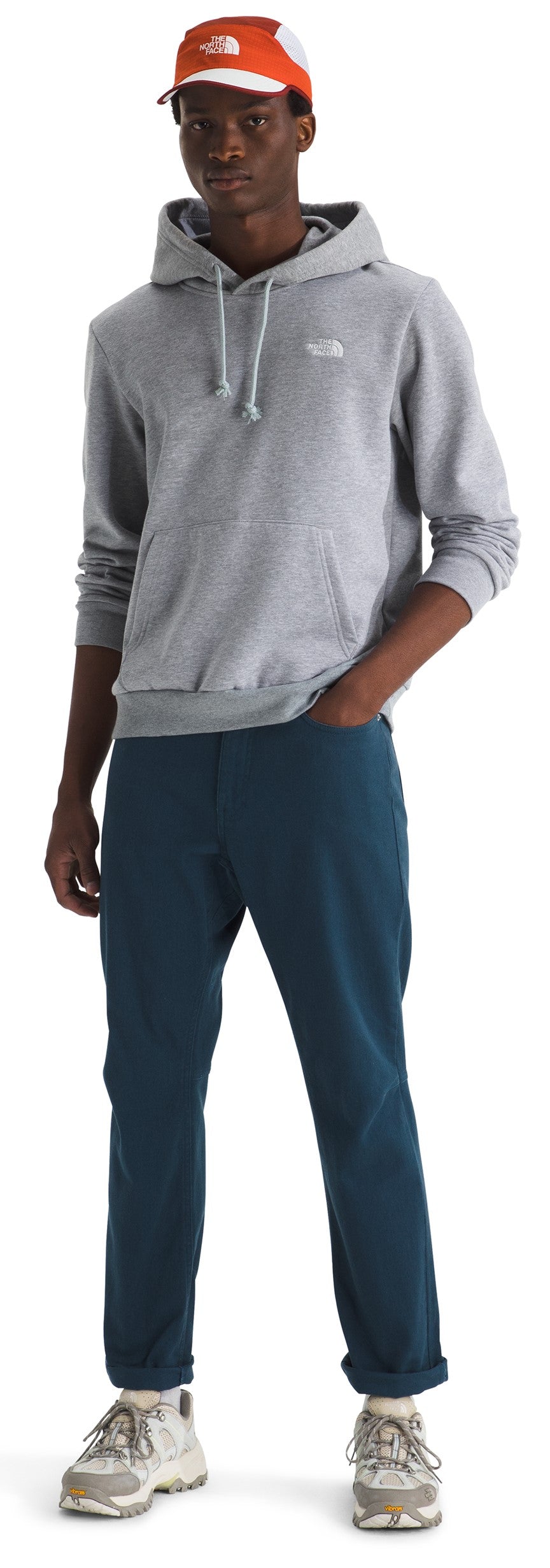 A male model stands wearing a light grey hooded sweatshirt with a small white logo on the chest, dark teal pants, a white and orange baseball cap, and beige athletic shoes.