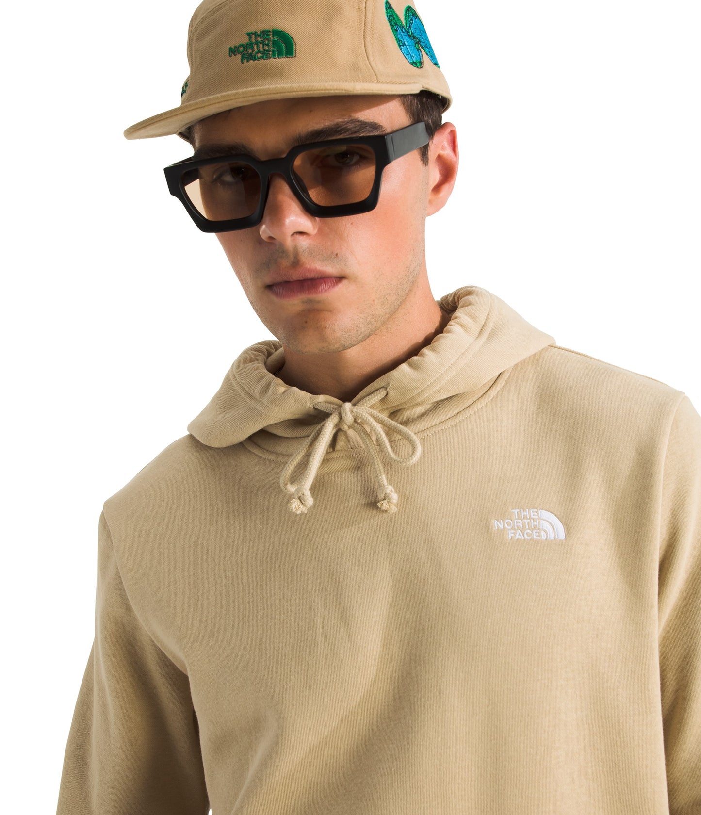 Man wearing beige hoodie with white logo, tan cap with green logo and butterfly, and black glasses.