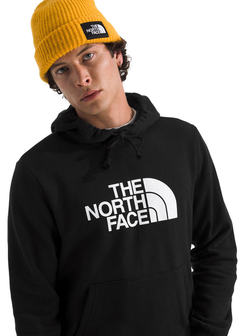 A man wearing a yellow ribbed knit beanie with a square patch logo on the front and a black hooded sweatshirt with a large white The North Face logo printed across the chest.