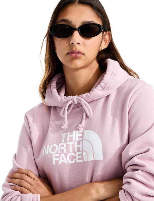 A young woman wearing a light pink hooded sweatshirt with the words THE NORTH FACE printed in large white letters across the chest, paired with black oval-shaped sunglasses.
