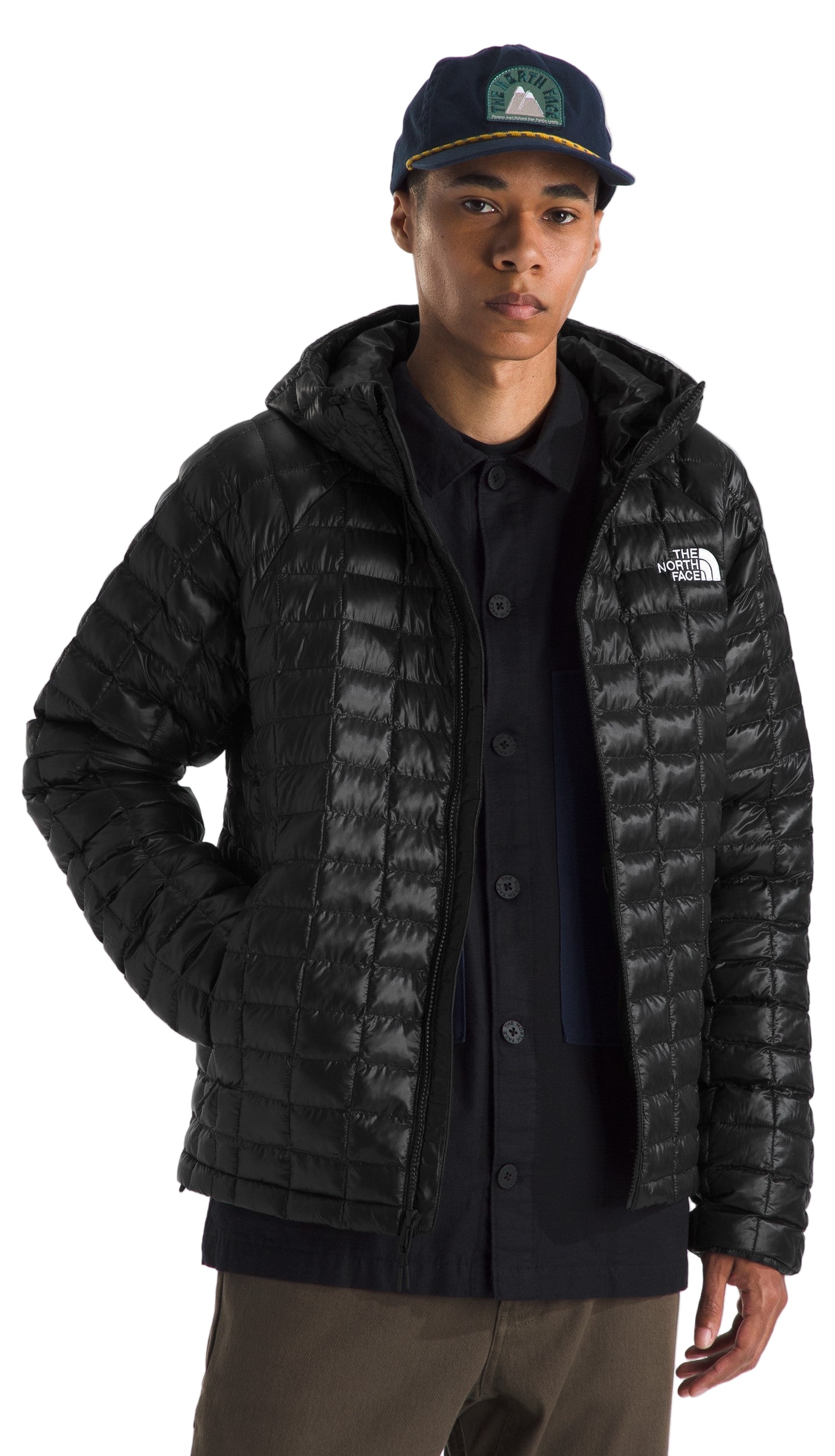 A person wearing a black quilted jacket with a hood, a black button-down shirt, and a dark blue hat.