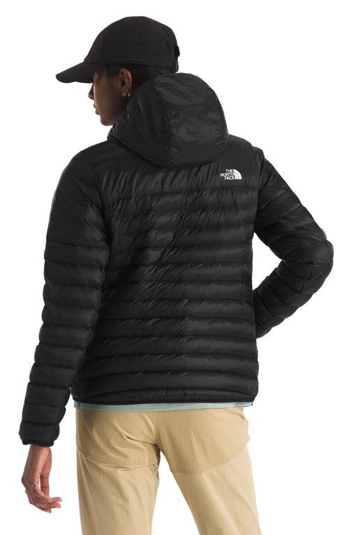 A Black hooded down jacket on a model with khaki pants and a black baseball cap. The jacket has horizontal stitching patterns and a white logo on the upper right side.