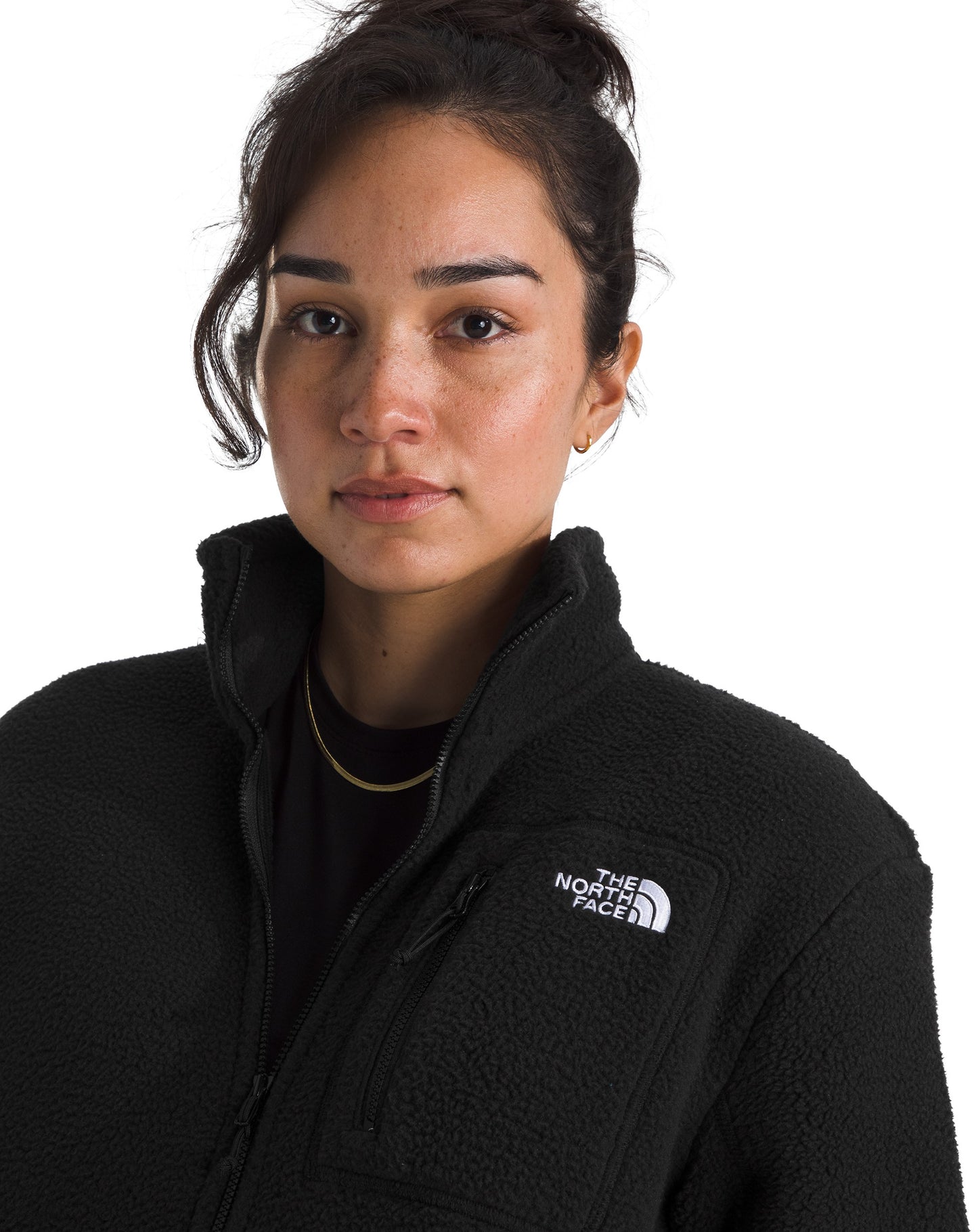 A woman with her dark hair pulled up wears a black fleece jacket with a high collar and a zippered chest pocket that features the white embroidered text THE NORTH FACE.