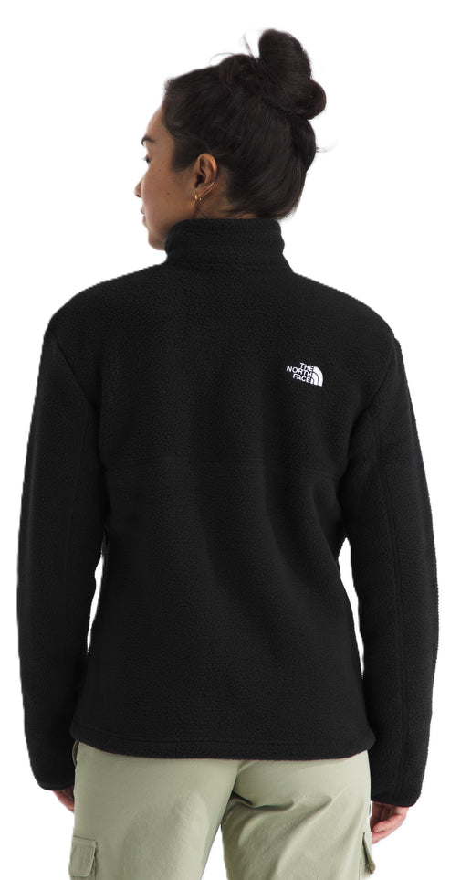 Rear view of a person with dark hair in a bun wearing a black fleece zip-up jacket with a white logo on the back right shoulder and light green cargo pants.