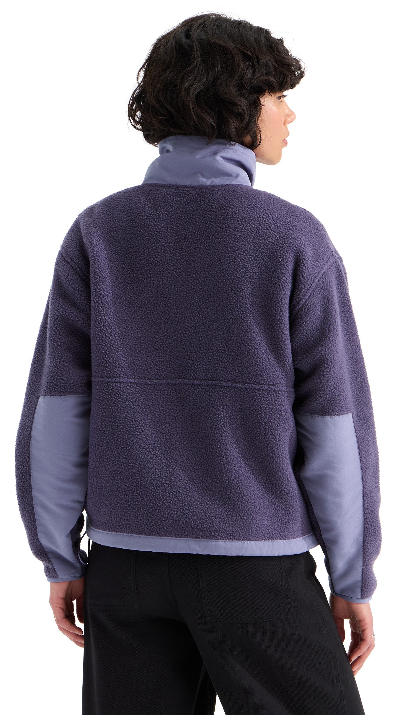 A back view of a person with short, wavy dark hair wearing a long-sleeved, high-neck fleece jacket in a muted dark purple color, featuring lighter purple panels on the lower sleeves and black pants.