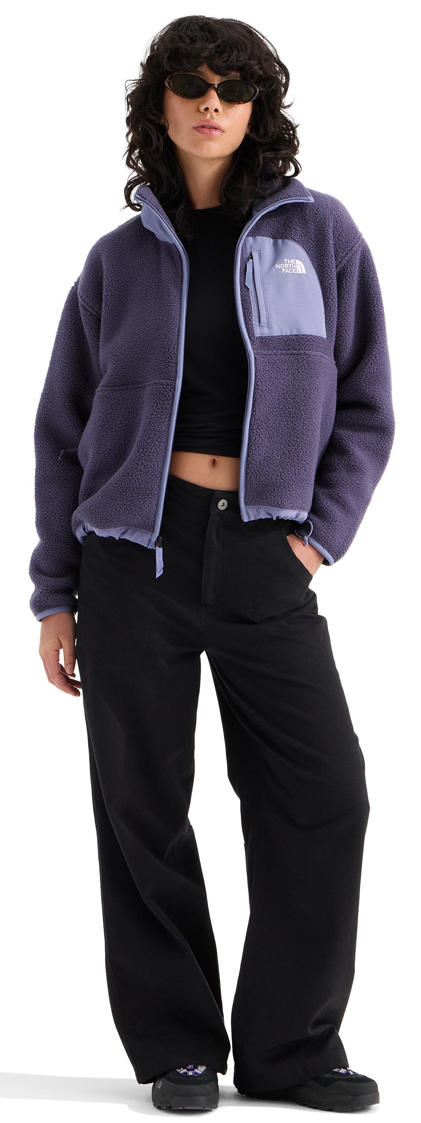 A person wearing a purple zip-up fleece jacket with a chest pocket, a black cropped shirt, and loose black trousers, standing with one hand in their pocket while wearing dark sunglasses.