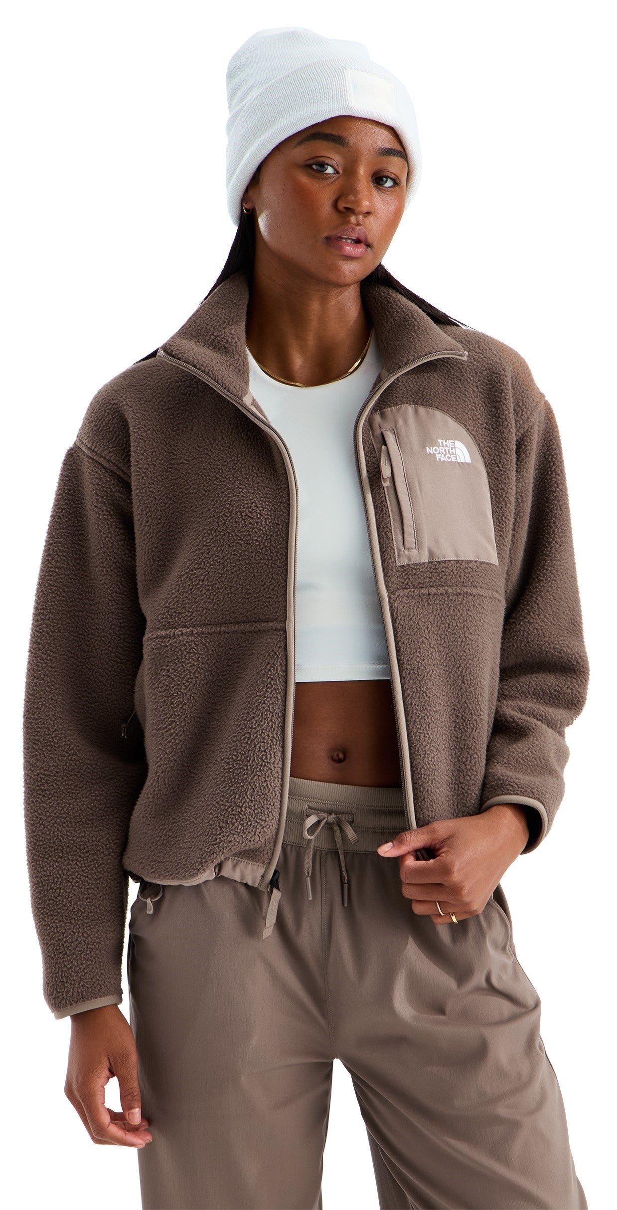 A woman wearing a brown fuzzy zip-up fleece jacket with a small zippered chest pocket, a white beanie, and a white crop top, paired with brown relaxed-fit pants.