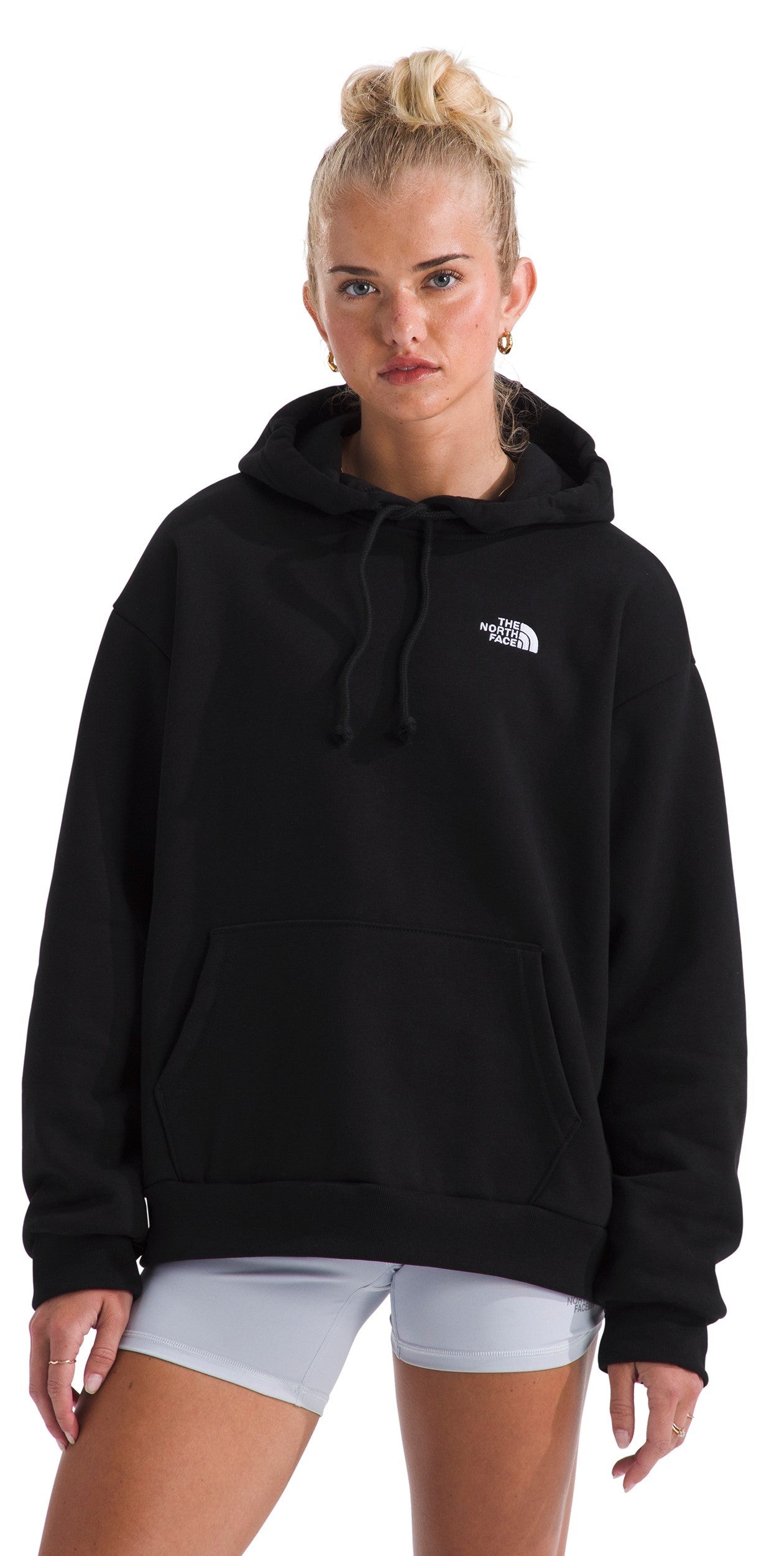A woman wearing a black, long-sleeved hooded sweatshirt with a small white logo on the left chest, paired with light gray fitted shorts.