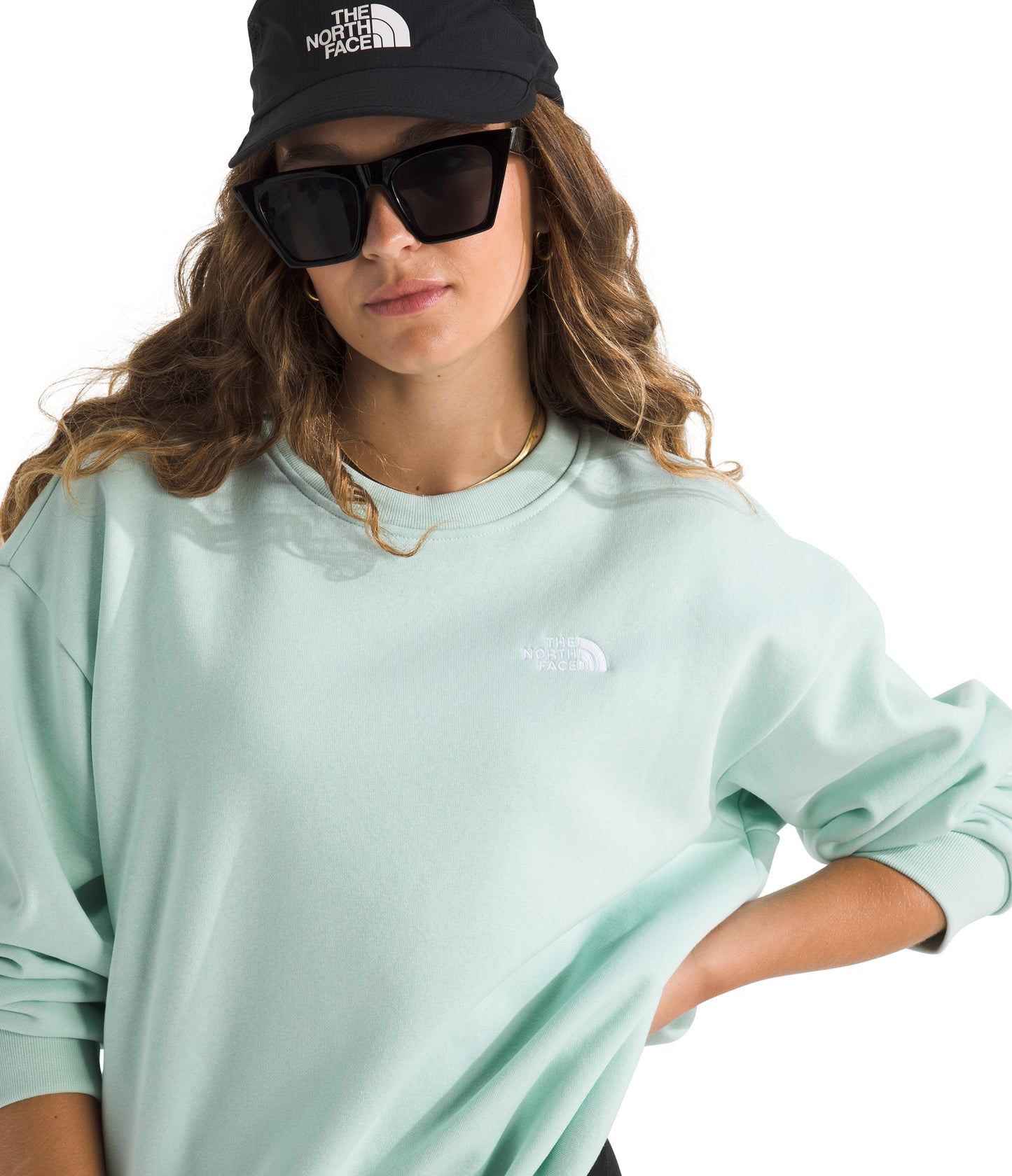A woman wearing a light-green crew neck sweatshirt, black baseball cap with white text, and black sunglasses.