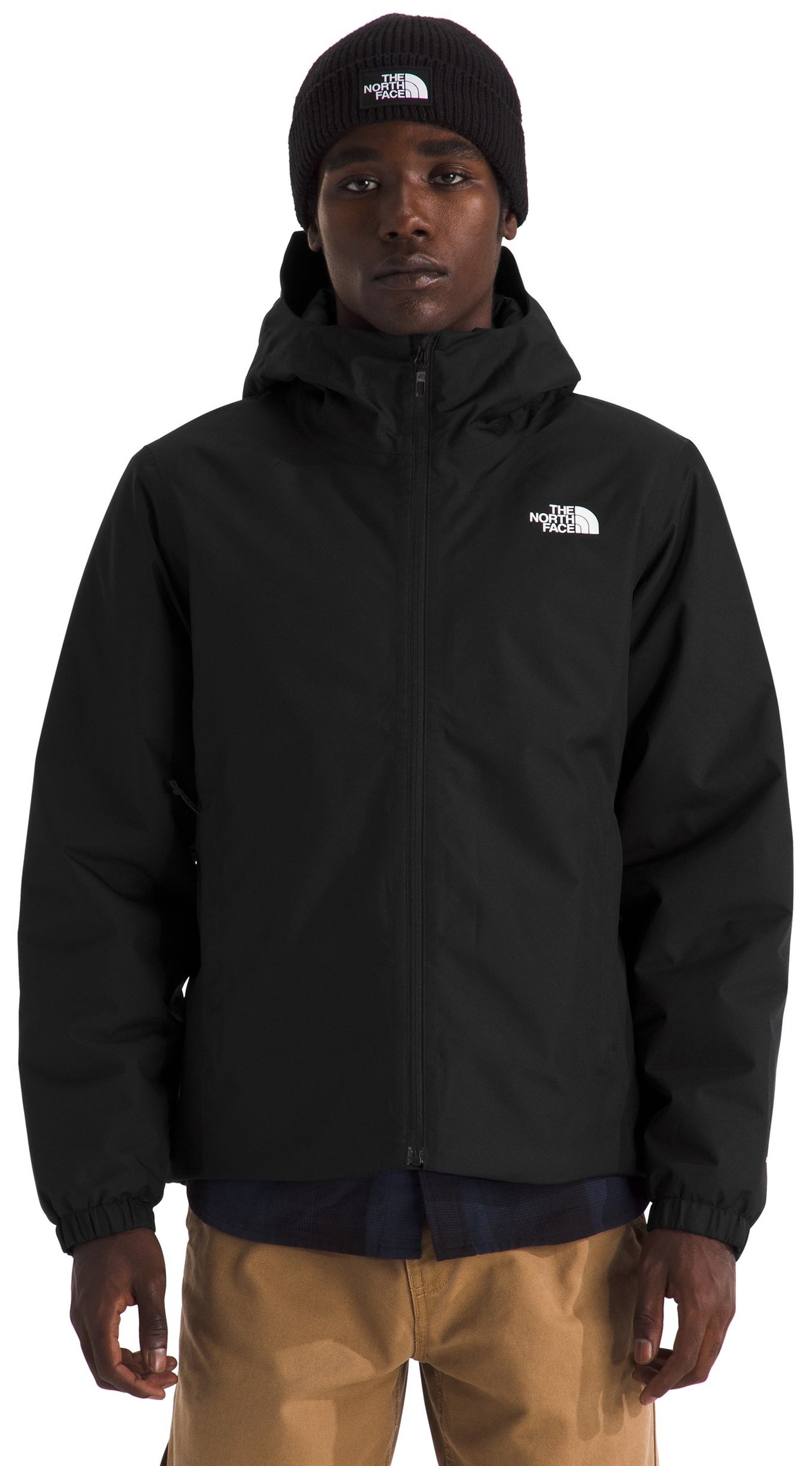 A male model wearing a black, hooded, zip-up jacket with a matching black beanie, both featuring a white logo, over a dark shirt and khaki pants.