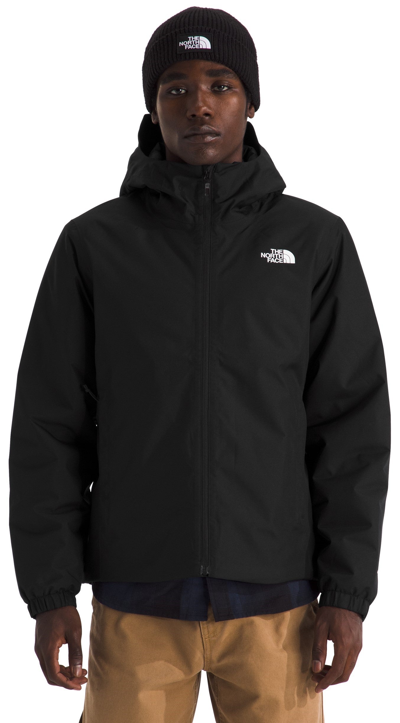 A male model wearing a black, hooded, zip-up jacket with a matching black beanie, both featuring a white logo, over a dark shirt and khaki pants.