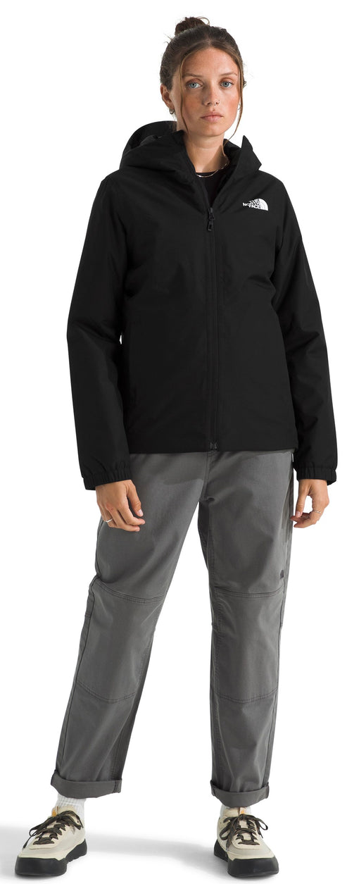 A woman stands facing forward, wearing a black hooded zip-up rain jacket with a small white logo on the left chest, grey trousers with cuffed hems, and light-colored sneakers.