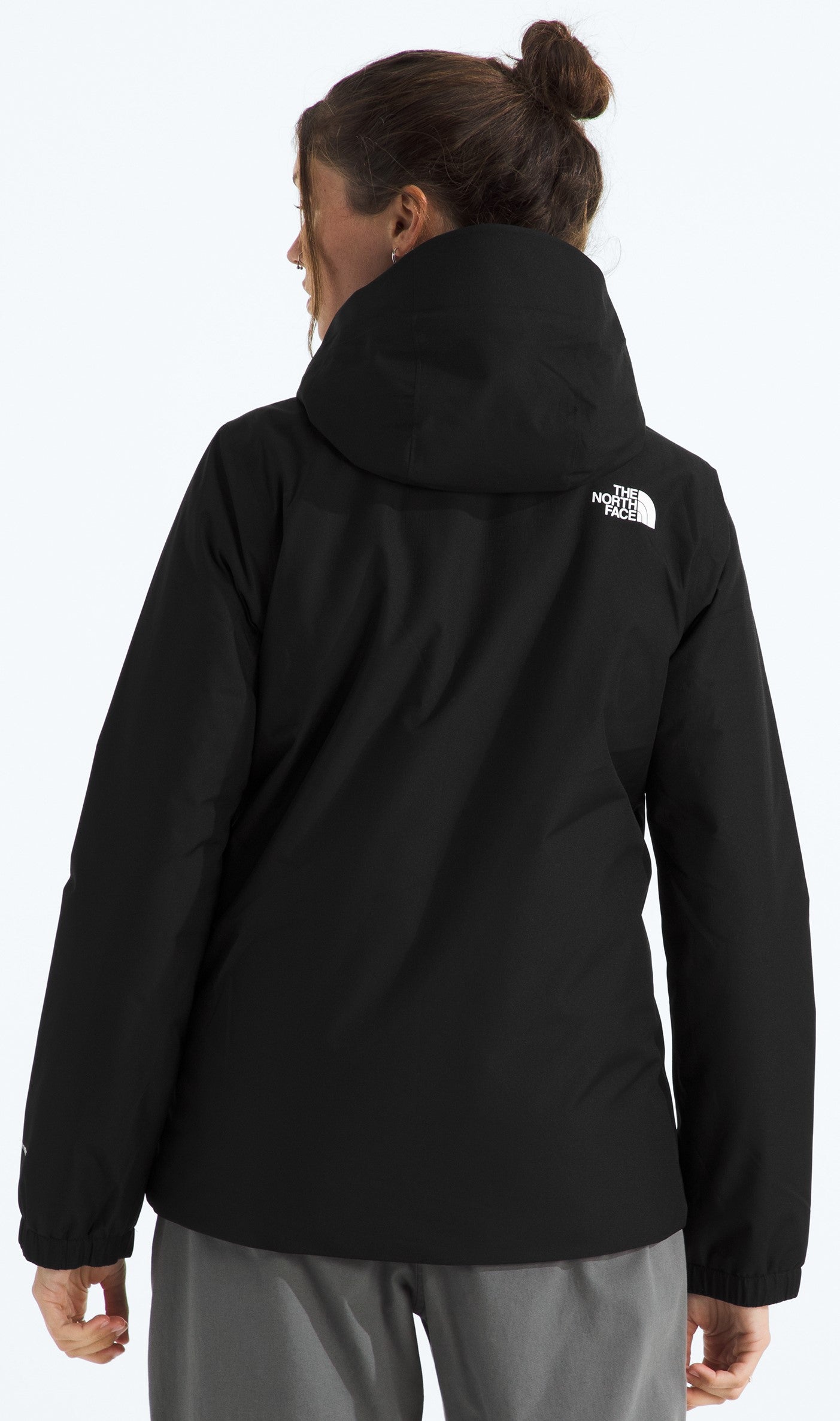 A back view of a woman wearing a black, hooded, long-sleeved jacket with a white logo on the back right shoulder, paired with grey pants.
