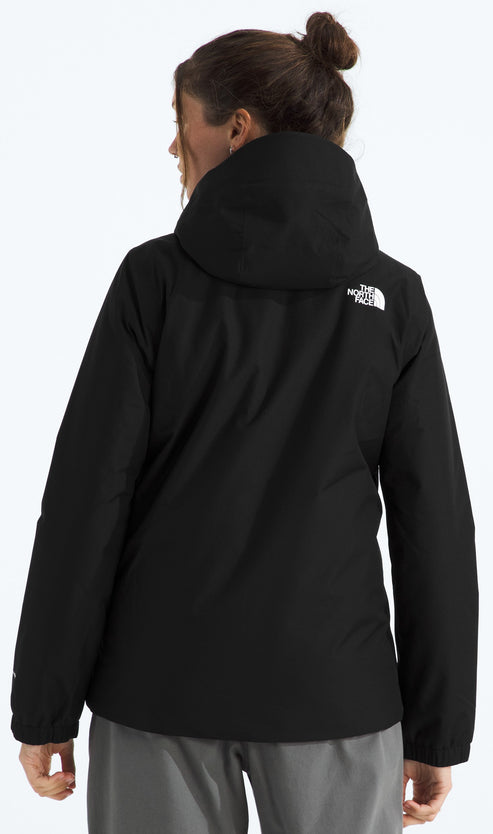 A back view of a woman wearing a black, hooded, long-sleeved jacket with a white logo on the back right shoulder, paired with grey pants.