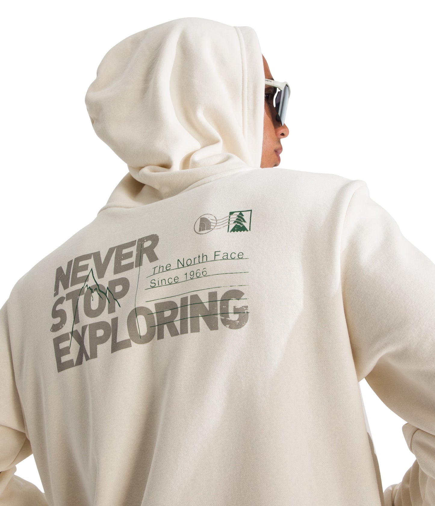 A person wears a cream-colored hoodie. On the back, in dark lettering, it says "NEVER STOP EXPLORING," with a mountain line drawing incorporated into the first two words. To the right of the text are horizontal lines representing a mailing address format, with a small postage-style stamp graphic featuring a pine tree above the lines. Small text reads "The North Face" and "Since 1966."