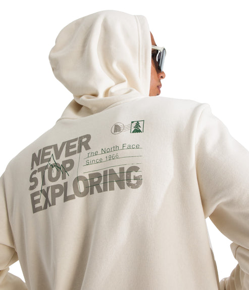 A person wears a cream-colored hoodie. On the back, in dark lettering, it says "NEVER STOP EXPLORING," with a mountain line drawing incorporated into the first two words. To the right of the text are horizontal lines representing a mailing address format, with a small postage-style stamp graphic featuring a pine tree above the lines. Small text reads "The North Face" and "Since 1966."