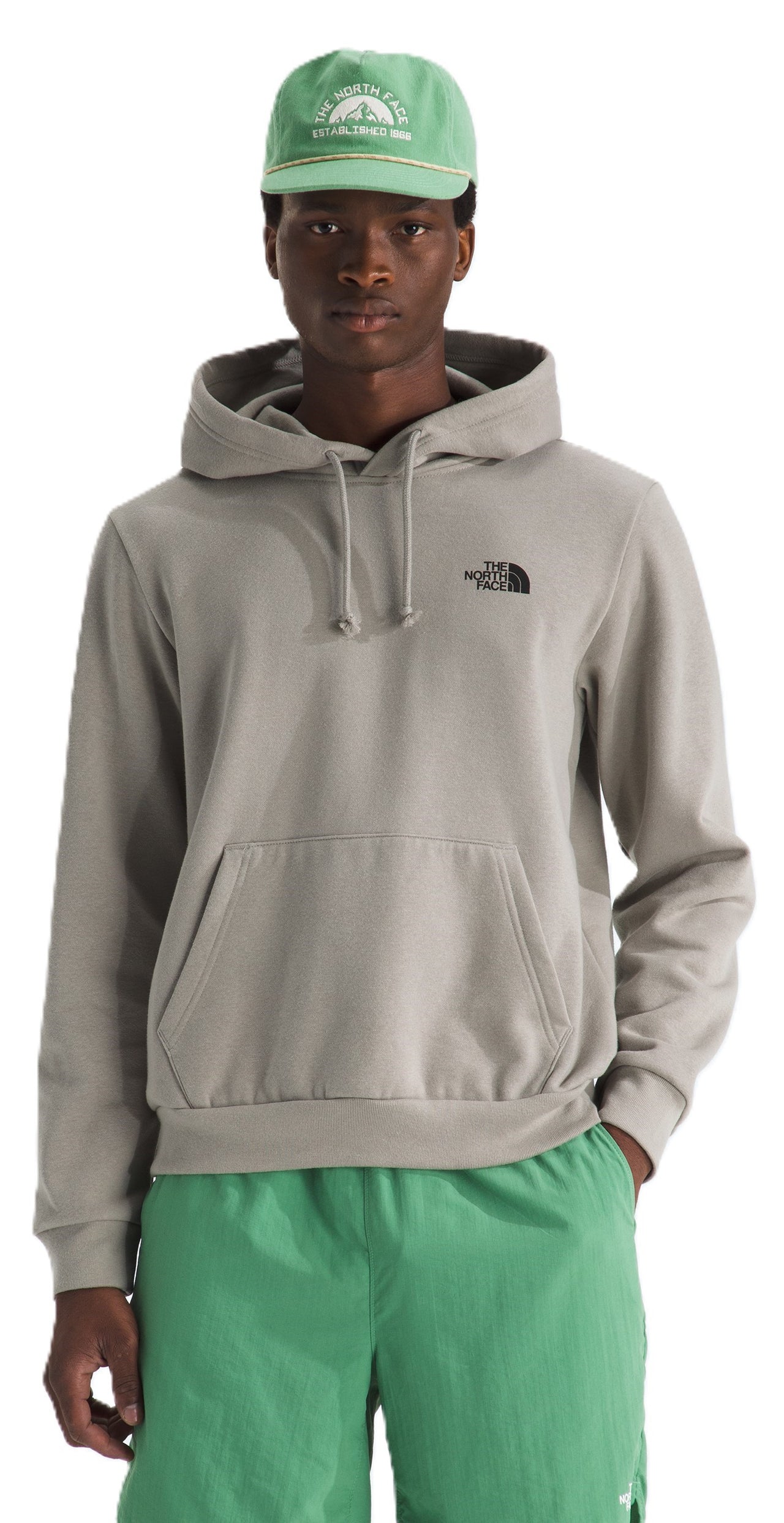 A man wearing a grey hooded sweatshirt with a small black logo on the left chest, a green baseball cap with a graphic on the front, and green shorts.