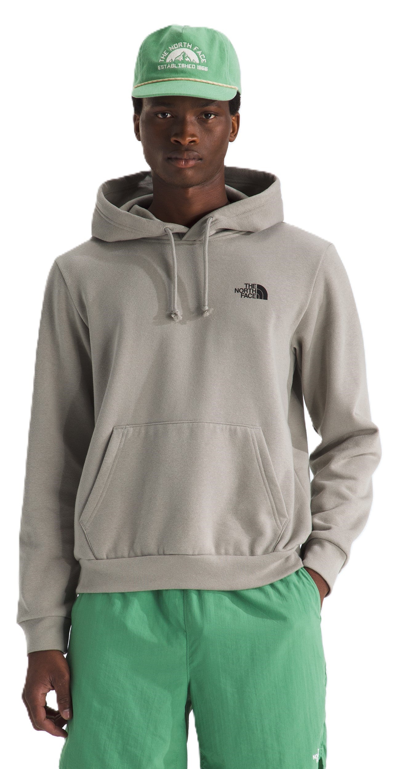 A man wearing a grey hooded sweatshirt with a small black logo on the left chest, a green baseball cap with a graphic on the front, and green shorts.