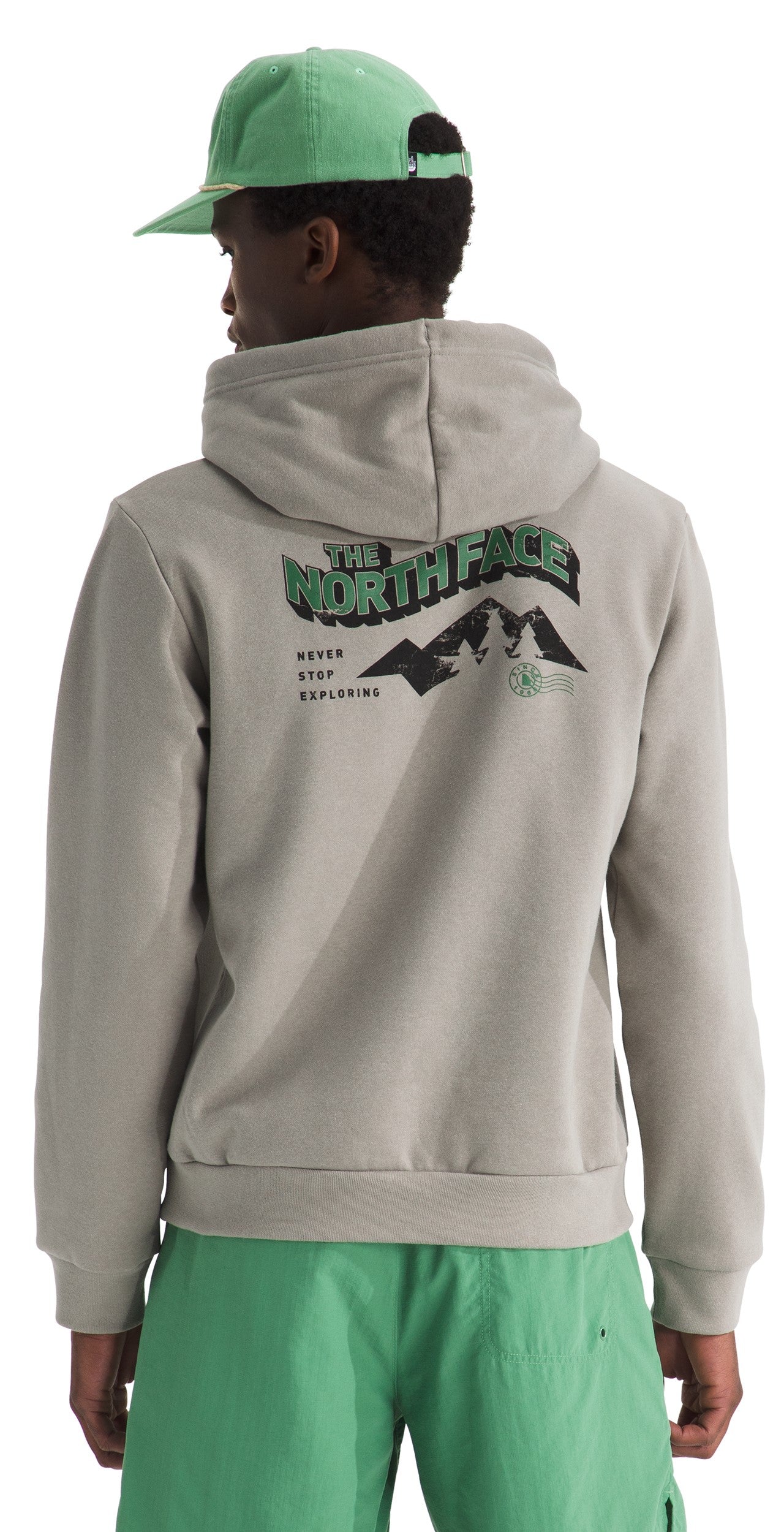 A person viewed from behind wearing a light beige pullover hoodie with a hood and a green baseball cap, with the back of the hoodie featuring a graphic print of a mountain range, text that reads THE NORTH FACE and NEVER STOP EXPLORING, and wearing bright green shorts.