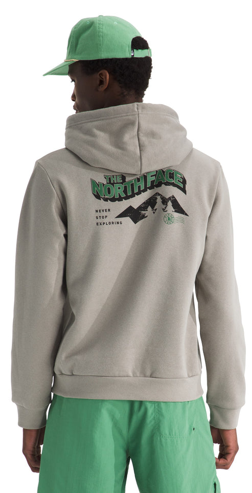 A person viewed from behind wearing a light beige pullover hoodie with a hood and a green baseball cap, with the back of the hoodie featuring a graphic print of a mountain range, text that reads THE NORTH FACE and NEVER STOP EXPLORING, and wearing bright green shorts.
