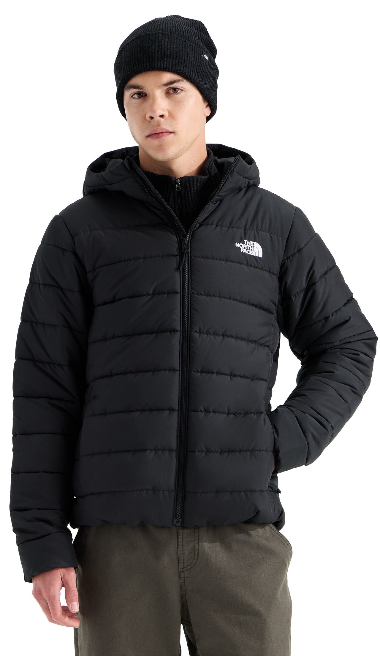 A man wearing a black hooded puffer jacket, a black beanie, and khaki pants against a white background.