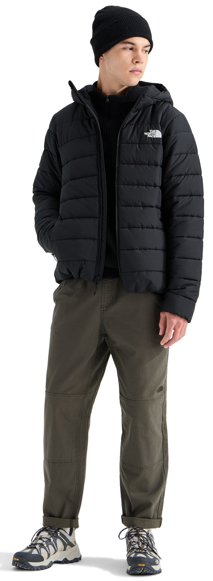 A male model standing wearing a black beanie, a black hooded puffer jacket, a black mock-neck shirt underneath, olive green trousers, and gray hiking sneakers.