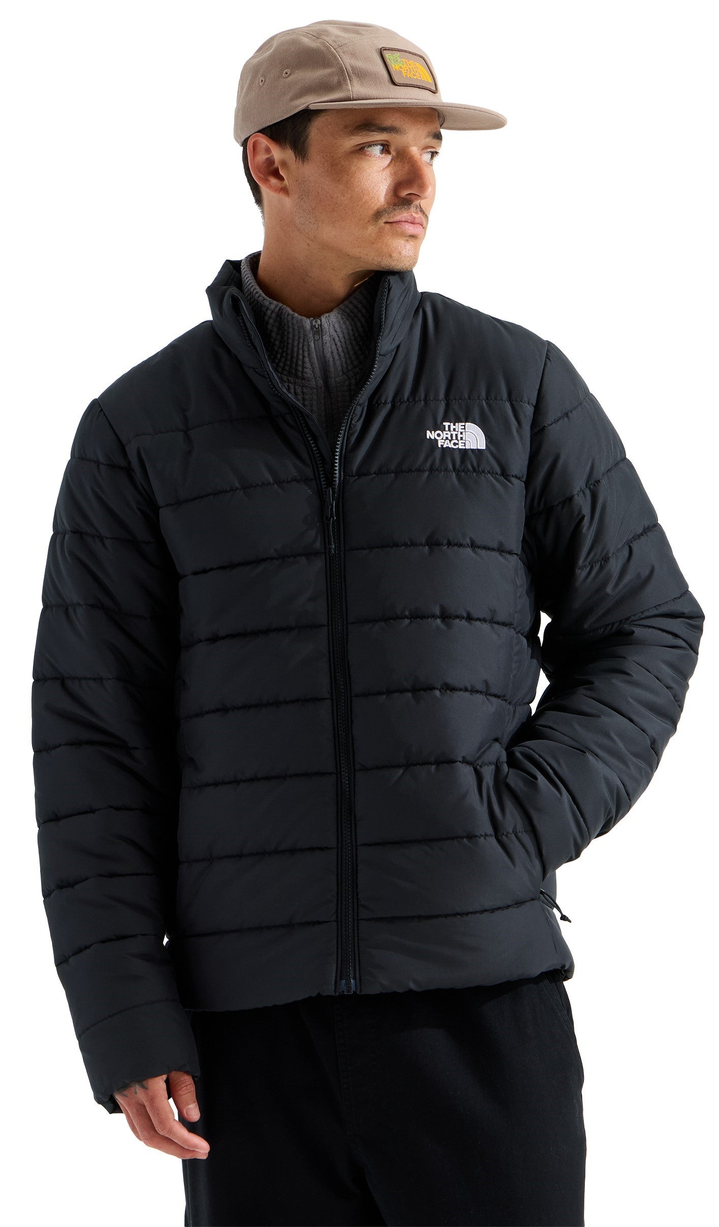A man wearing a tan baseball cap with a square patch, a grey mock-neck sweater, and a black puffer jacket with horizontal quilting and the white The North Face logo on the chest.