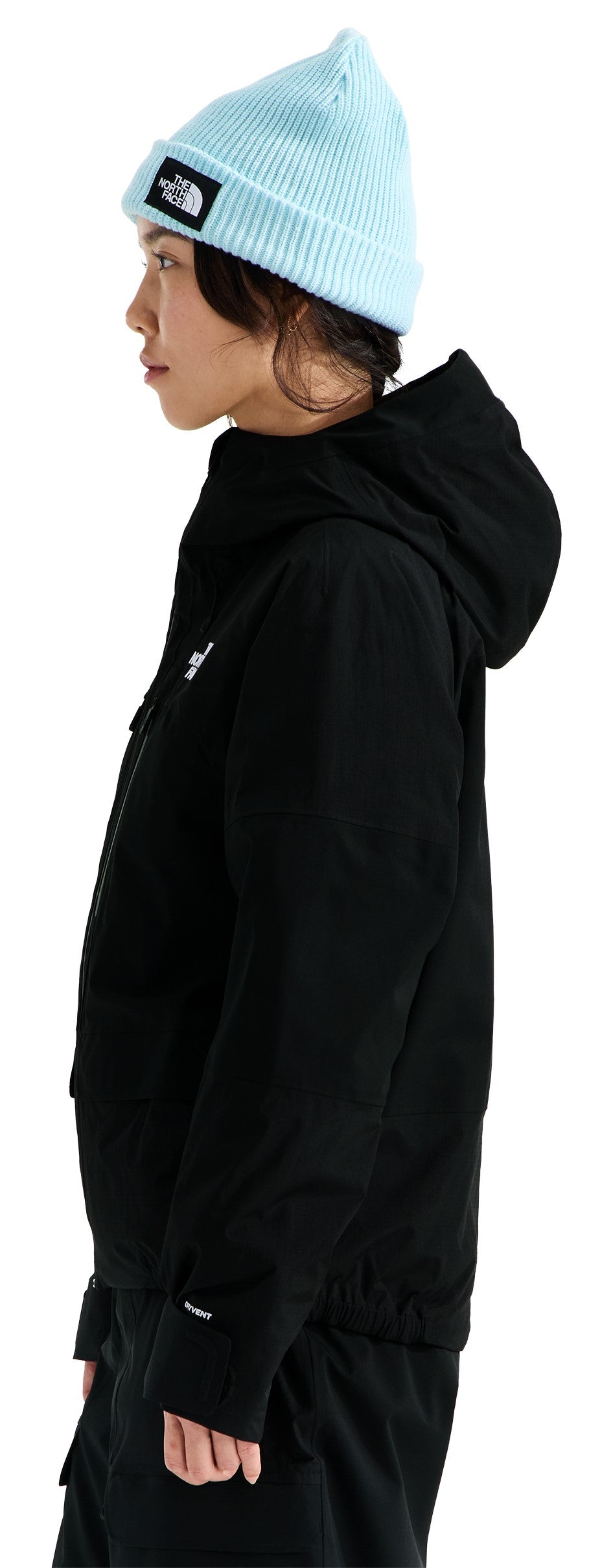A person viewed from the side wearing a light blue ribbed beanie with a black and white rectangular patch on the front, and a black, long-sleeved, hooded jacket featuring a small white logo on the upper chest.