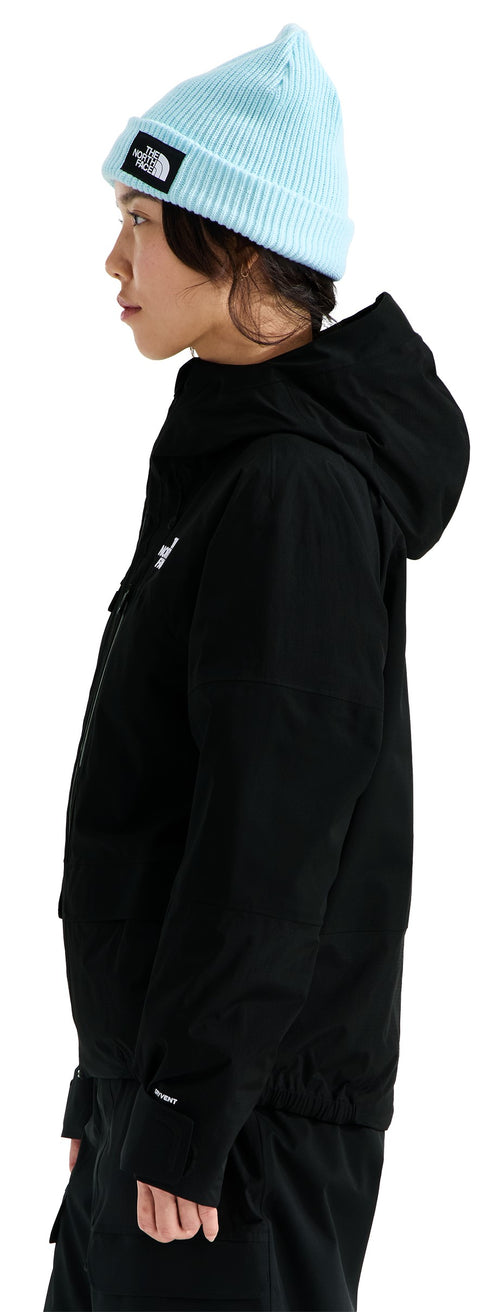 A person viewed from the side wearing a light blue ribbed beanie with a black and white rectangular patch on the front, and a black, long-sleeved, hooded jacket featuring a small white logo on the upper chest.