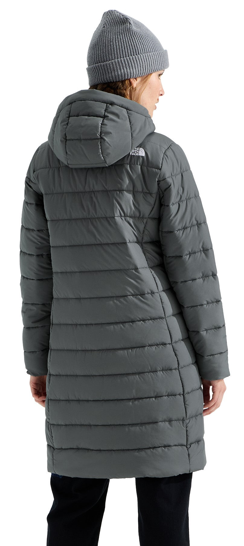 A back view of a woman wearing a charcoal gray, knee-length, quilted puffer coat with a hood and a matching charcoal gray knit beanie, seen against a white background.
