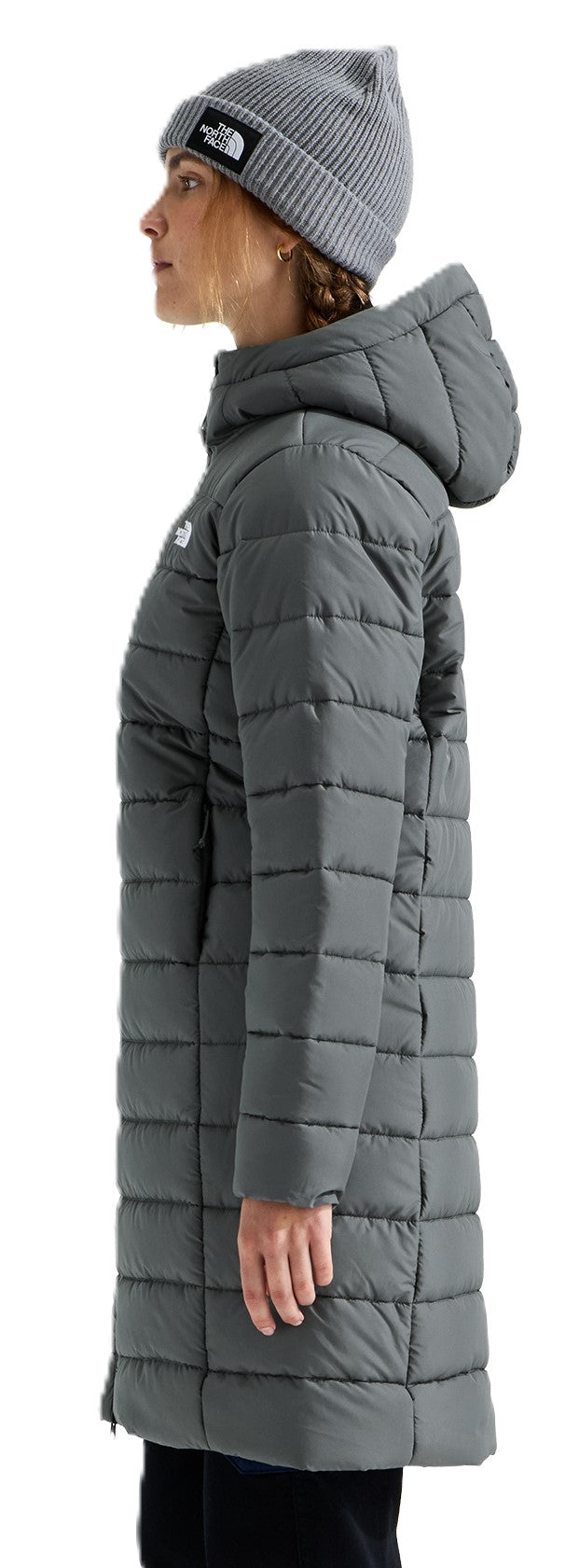 A woman wearing a gray, hooded, long puffer coat and a gray ribbed beanie cap, viewed from the side.
