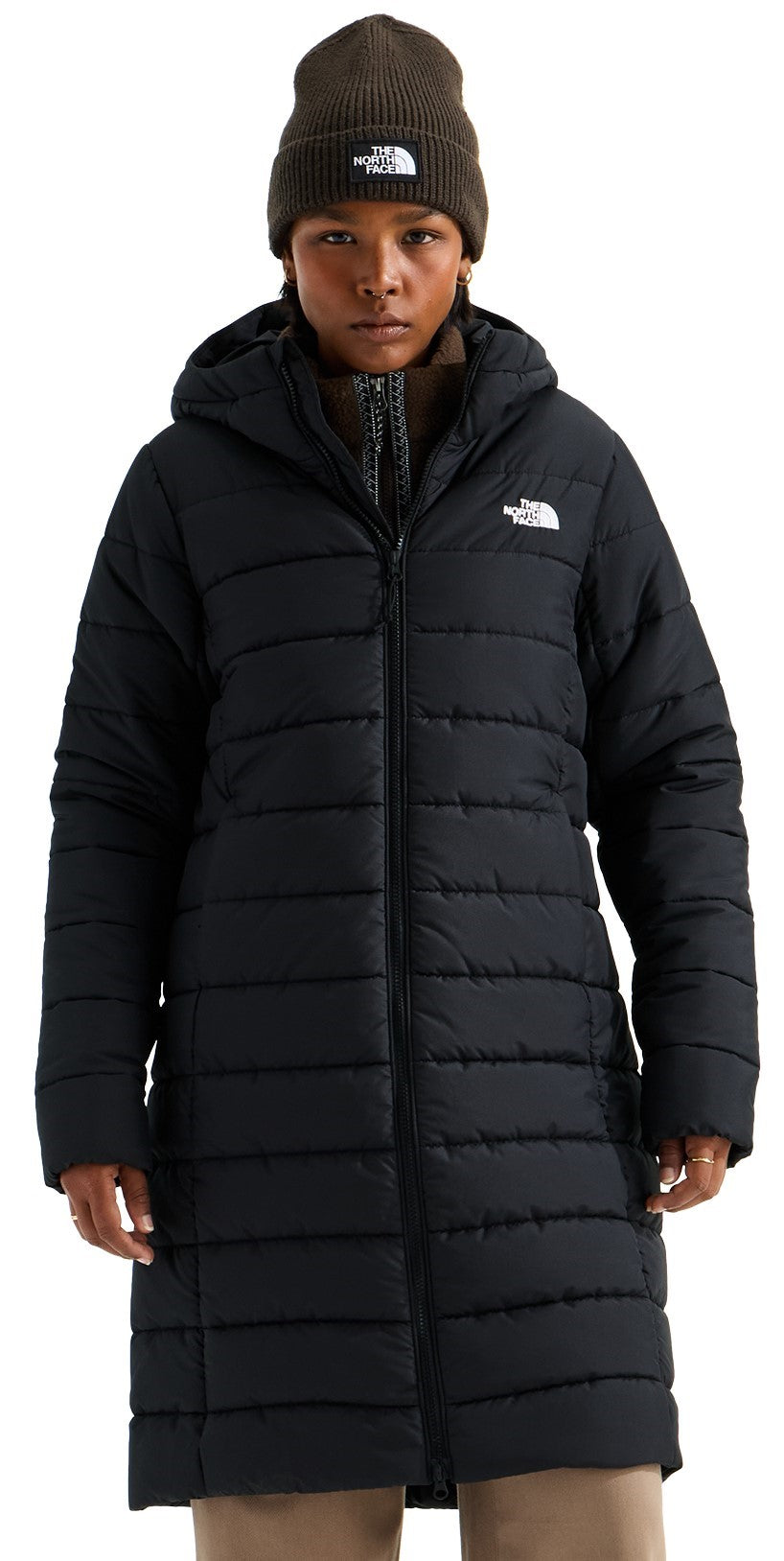 A person wearing a black, knee-length, hooded, quilted puffer coat with a front zipper closure and a dark olive beanie featuring a logo on the front.