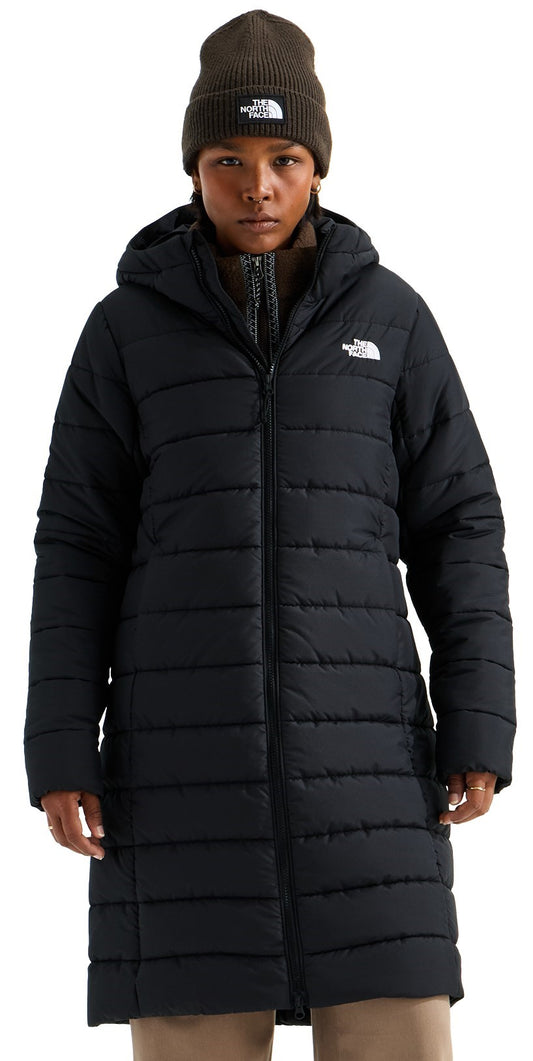 A person wearing a black, knee-length, hooded, quilted puffer coat with a front zipper closure and a dark olive beanie featuring a logo on the front.