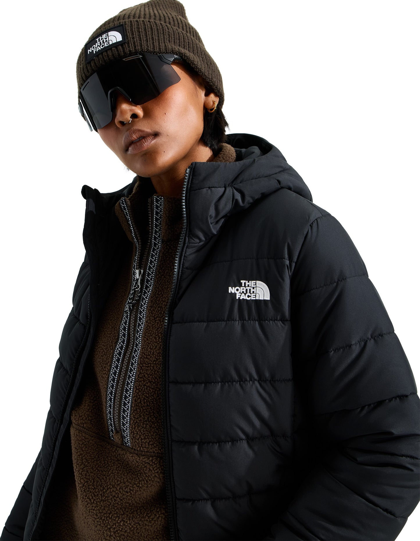 A person wearing a dark olive beanie with a rectangular white patch, large wraparound sports sunglasses, a brown fleece quarter-zip pullover, and a black hooded puffer jacket with The North Face logo on the chest.